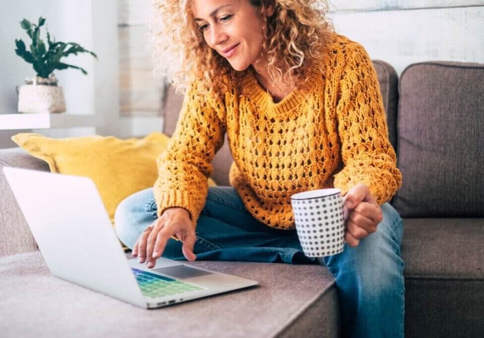 Woman with blonde curly hair sitting down on the sofa with lap top and coffee at home.